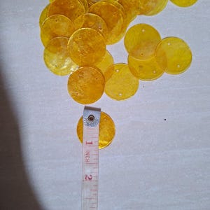 Könnte beinhalten: Eine Sammlung von durchscheinenden gelben runden Pailletten, die jeweils zwei kleine Löcher zur Befestigung aufweisen. Ein weißes Maßband liegt auf einer der Pailletten und zeigt einen Durchmesser von etwa 2,54 cm für jedes einzelne Stück an.