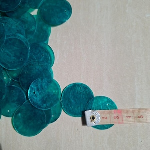 Peut inclure: Une collection de sequins circulaires et translucides de couleur sarcelle, chacun doté d'un petit trou près du bord supérieur. Un ruban à mesurer est placé sur l'un des sequins, indiquant un diamètre d'environ 3,2 cm pour chaque pièce.