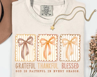 May include: Cream-colored Comfort Colors XL t-shirt with the words "Grateful, Thankful, Blessed" and "God is faithful in every season." Three watercolor bows are featured. A gold star pendant necklace is draped across the shirt.