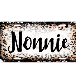 Puede incluir: Placa de matrícula rectangular con un borde de estampado de leopardo en tonos marrón, negro y blanco. La palabra "Nonnie" está escrita en una fuente cursiva grande y negra en el centro de la placa.