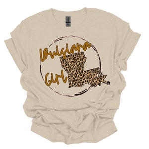 May include: A beige t-shirt with a brown circle design featuring the outline of Louisiana in a leopard print pattern and the text "Louisiana Girl".