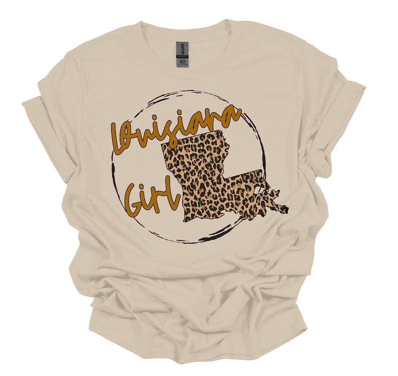 May include: A beige t-shirt with a brown circle design featuring the outline of Louisiana in a leopard print pattern and the text "Louisiana Girl".