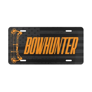 May include: Rectangular license plate with a black and gray American flag design. The plate features the word "BOWHUNTER" in orange block letters, along with an orange bow and arrow graphic on the left side. The plate has four mounting holes.