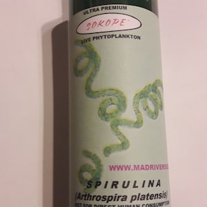 May include: A green glass bottle with a white cap, labeled "ULTRA PREMIUM SOKOPE LIVE PHYTOPLANKTON WWW.MADRIVERSE SPIRULINA (Arthrospira platensis) NOT FOR DIRECT HUMAN CONSUMPTION".