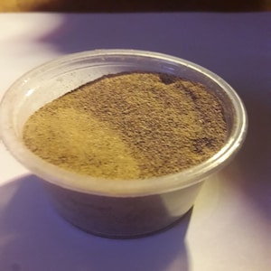 May include: A small plastic container filled with a light brown powder. The powder is a mixture of two shades of brown, with a lighter brown on the top and a darker brown on the bottom.