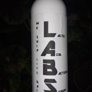 Op de afbeelding: Witte plastic fles met een zwart etiket waarop "LABS" staat, met de letters verticaal gestapeld. Het etiket heeft ook de tekst "WE SHIP LIFE DAILY" aan de linkerkant en "LACTIC ACID BACTERIA ERUM WWW.MA" aan de rechterkant.