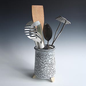 Könnte beinhalten: Ein Keramik-Utensilienhalter mit weiß-schwarz gesprenkelter Oberfläche und drei kleinen Füßen, der einen Holzspatel, einen geschlitzten Edelstahlwender, einen Metalllöffel und einen Kartoffelstampfer aus Draht aufrecht enthält.