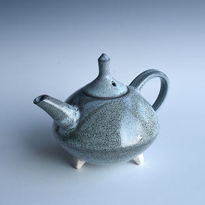 Könnte beinhalten: Eine gesprenkelte blaue Keramik-Teekanne mit rundem Körper, gebogenem Henkel und Ausguss. Der Deckel hat einen kleinen, spitzen Knopf. Die Teekanne steht auf drei kleinen, weißen Füßen. Das Design ist schlicht und elegant.
