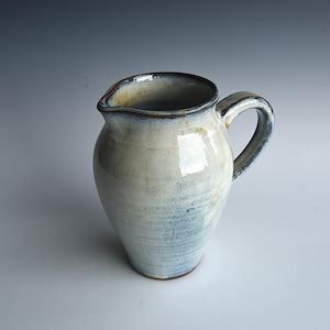 May include: A handmade ceramic pitcher with a rounded body and a curved handle. The pitcher is glazed in shades of white and blue, with a brown rim. The pitcher is a decorative item for the kitchen or dining room.