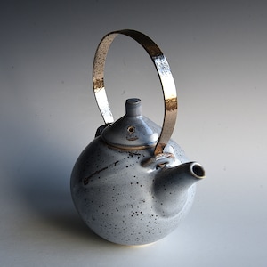 Könnte beinhalten: Eine Keramik-Teekanne mit einer gesprenkelten, hellblauen Glasur und einem gehämmerten Metallgriff. Die Teekanne hat einen abgerundeten Körper, einen kleinen Ausguss und einen Deckel mit einem kleinen Knopf. Der Griff ist gebogen und bogenförmig.