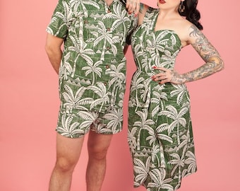 May include: A couple wearing matching green and white tropical print clothing. The man is wearing a short-sleeved shirt and matching shorts. The woman is wearing a sleeveless dress. Both are standing on a pink background.