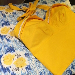 以下が含まれることがあります： 首元に白いトリムが付いた黄色のホルターネックトップ。トップは柔らかく軽量な生地で作られており、ゆったりとしたフィット感です。