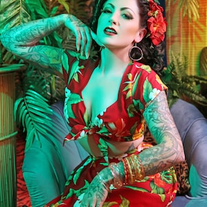 May include: A woman wearing a red and green tropical print crop top and matching skirt. She is sitting on a blue couch with a green plant behind her. She has a flower in her hair and is wearing bracelets on her wrist.