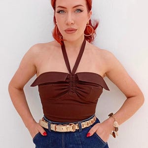 May include: A brown halter top with a gathered bodice and a matching halter strap. The top is paired with blue denim shorts and a gold-colored belt. The model is wearing gold hoop earrings and a bracelet with shell-like charms.