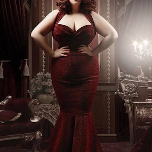 May include: A woman in a red, sparkly, mermaid-style dress poses in a luxurious setting. The dress has a sweetheart neckline and a fitted bodice. The woman has red hair and is wearing red lipstick.
