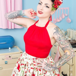 Könnte beinhalten: Eine Frau mit einem roten Halstuch und einem weißen Rock mit einem roten und grünen Küchen-Motiv. Sie trägt eine rote Blume im Haar und rote Ohrringe. Der Hintergrund ist eine blaue und weiße Küche.