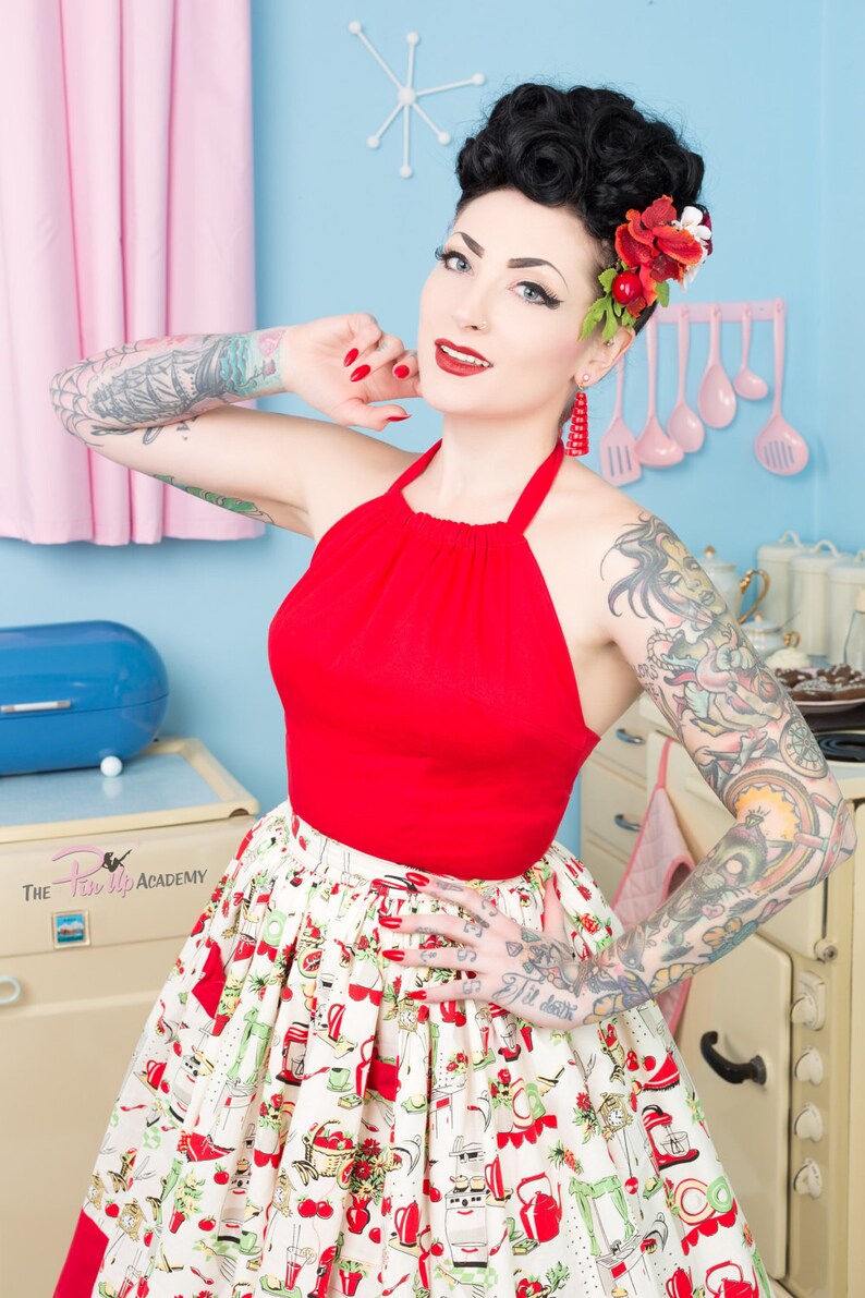 Może przedstawiać: Kobieta w czerwonym topie na ramiączkach i białej sp&oacute;dnicy z czerwono-zielonym nadrukiem w stylu kuchennym. Ma czerwony kwiat we włosach i czerwone kolczyki. Tło to niebieska i biała kuchnia.