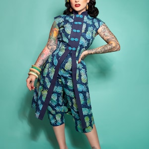 May include: A woman wearing a blue and green pineapple print dress with a mandarin collar and a blue sash. She is wearing a flower in her hair and has a tattoo on her arm.