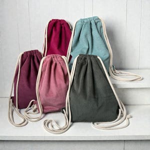 Könnte beinhalten: Fünf Kordelzugbeutel aus Cord in Rosa-, Türkis- und Grüntönen. Jeder Beutel hat einen passenden Kordelzugverschluss und weiße Seilriemen. Die Beutel sind auf einer weißen Oberfläche vor einem weißen Hintergrund angeordnet.