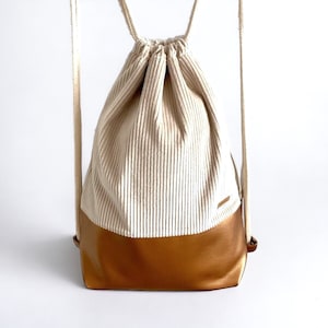 Könnte beinhalten: Ein weißer und brauner Cord-Rucksack mit Kordelzug und goldfarbenem Lederboden.