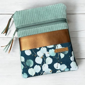 Könnte beinhalten: Eine tealblaue Reißverschlusstasche mit einem goldfarbenen Kunstlederakzent und einem weißen und tealfarbenen Blumendruck. Die Tasche hat einen braunen Quastenanhänger und der Text "kodanin" ist auf der Vorderseite aufgedruckt.