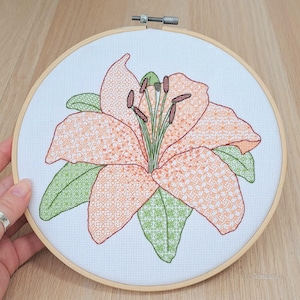 May include: A cross-stitch embroidery of a lily flower in a wooden hoop. The flower has orange petals with a geometric pattern, green leaves, and dark brown stamens. The background is white fabric.
