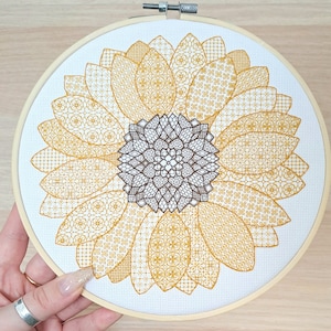 Op de afbeelding: Een close-up van een voltooide borduurring met een zonnebloemontwerp. De zonnebloem is gemaakt van meerdere lagen gele en bruine steken, wat een gedetailleerd en ingewikkeld patroon creëert.