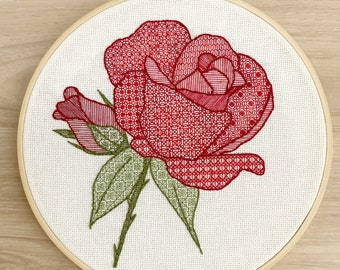 May include: A red embroidered rose with green leaves on a white background. The rose is stitched with a variety of patterns and is displayed in a wooden embroidery hoop.