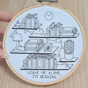 Könnte beinhalten: Schwarz-weißer Kreuzstich-Stickrahmen mit einem Bücherregal-Design mit Büchern, einer Lampe, einem Kaktus und dem Text „LEAVE ME ALONE, I'M READING“. Die Stickerei ist in einem Holzrahmen gefasst.