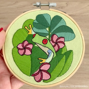 May include: Embroidered hoop art featuring a green tree frog perched on a leafy branch with pink flowers. The frog has a red eye and is surrounded by green foliage.