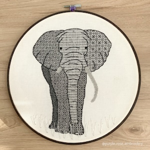 May include: Black and white embroidered elephant design on a white canvas in a wooden embroidery hoop. The elephant is standing with its trunk raised and has a detailed pattern on its body.