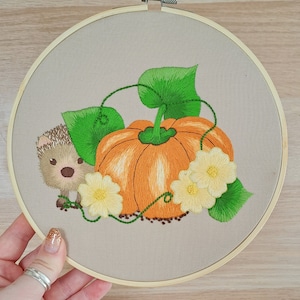 May include: Embroidered hoop art featuring a brown hedgehog next to a large orange pumpkin with green leaves and white flowers. The background is a beige fabric.