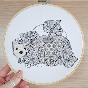 May include: A cross-stitch embroidery hoop featuring a hedgehog nestled beside a pumpkin and leaves. The design is in black and white, with intricate patterns filling the shapes. The embroidery is set against a white fabric background, held within a wooden hoop.