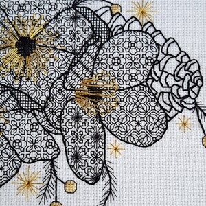 May include: Close-up of a cross-stitch embroidery featuring a floral design. The artwork showcases black and gold thread on a white background. The floral elements have intricate geometric patterns, with gold accents and starburst details.