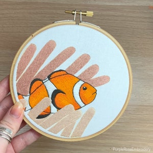 May include: Embroidered hoop art featuring an orange and white clownfish swimming amongst coral-like shapes in shades of peach and beige.