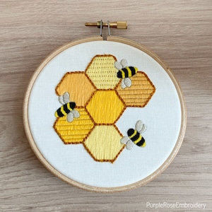 May include: Embroidered artwork of a honeycomb with three bees. The honeycomb is made up of yellow and orange hexagons. The bees are black and yellow with grey wings. The embroidery is set in a wooden hoop.
