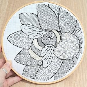May include: A cross-stitch embroidery of a bee and flower design. The bee is in the center, surrounded by petals with intricate geometric patterns. The embroidery is in black and white, set in a wooden hoop. The text "PurpleRoseEmbroidery" is in the upper left corner.