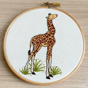 May include: An embroidered giraffe in a wooden hoop. The giraffe features a brown and tan spotted pattern, with green grass at its feet. The embroidery is on white fabric, and the hoop is secured with a brass screw.