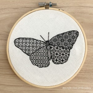 May include: Black and white embroidery of a butterfly with intricate geometric patterns on its wings, displayed in a wooden embroidery hoop.