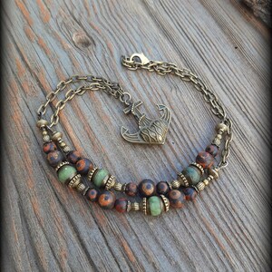 May include: A double-strand bracelet featuring bronze-toned metal chains, an anchor-shaped charm, and a series of round beads in mottled brown and solid light green. The bracelet includes decorative metal spacers and a lobster claw clasp.