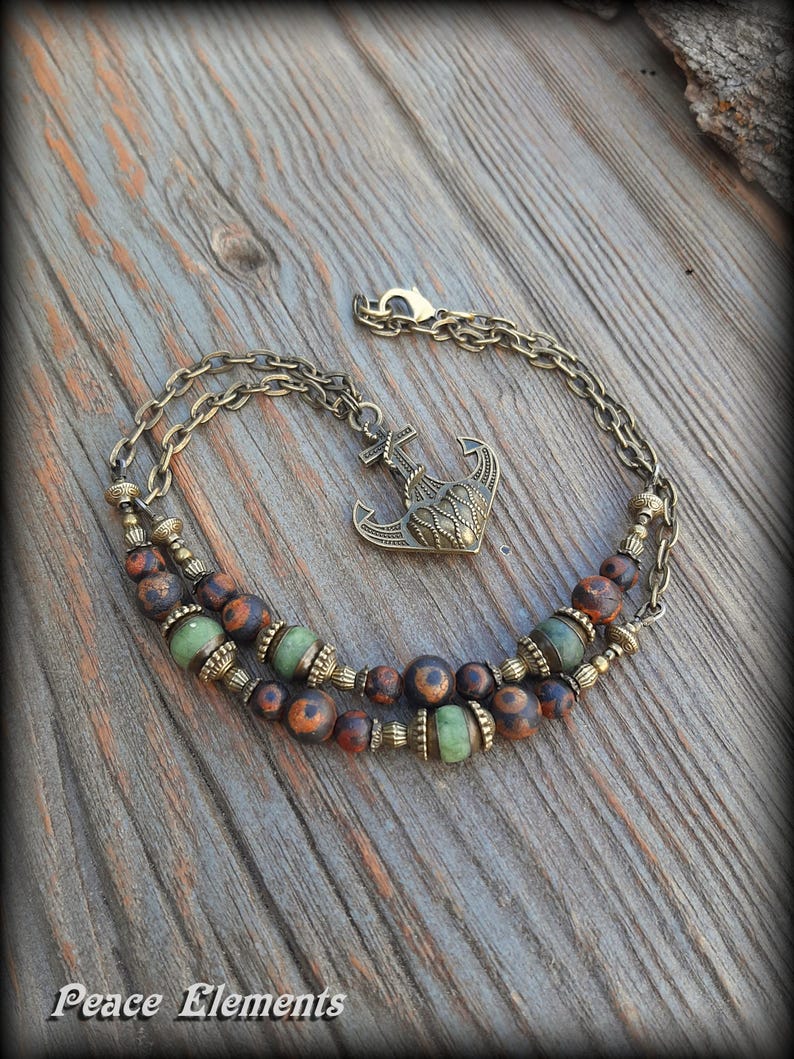 May include: A double-strand bracelet featuring bronze-toned metal chains, an anchor-shaped charm, and a series of round beads in mottled brown and solid light green. The bracelet includes decorative metal spacers and a lobster claw clasp.