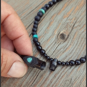 Puede incluir: Un collar de cuentas negras con un pequeño colgante de piedra turquesa. El colgante es un pequeño recipiente cilíndrico que se puede abrir y cerrar.