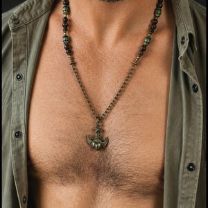 May include: A necklace featuring a bronze-toned anchor pendant suspended from a chain. The upper portion of the necklace consists of dark brown wooden beads interspersed with smaller, metallic green beads and bronze-colored spacers.