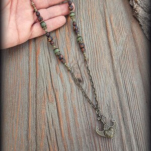 May include: A necklace featuring a bronze-toned metal chain with alternating matte brown and green beads. The pendant is a bronze-toned metal anchor with a heart-shaped center. The text Peace Elements is visible in the bottom left corner.