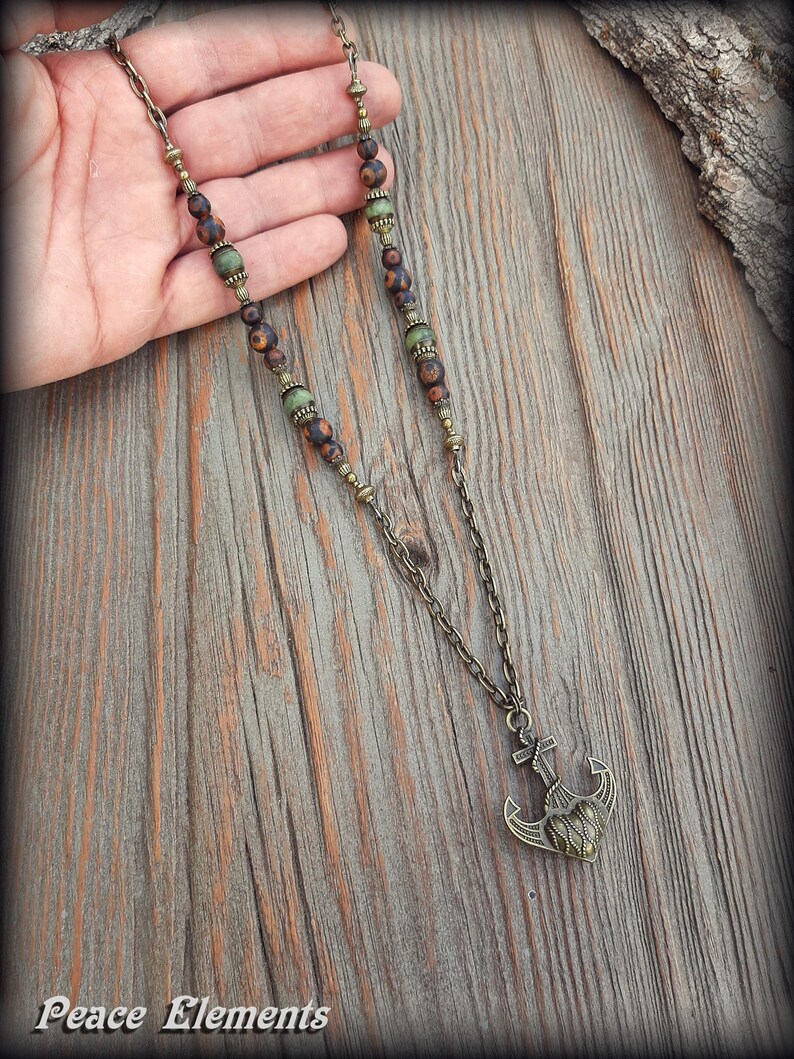 May include: A necklace featuring a bronze-toned metal chain with alternating matte brown and green beads. The pendant is a bronze-toned metal anchor with a heart-shaped center. The text Peace Elements is visible in the bottom left corner.