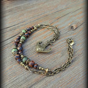 May include: A double-strand bracelet featuring bronze-toned metal chains and an anchor charm. The design incorporates round beads in mottled brown, blue, and green hues, accented with small, textured bronze-colored metal spacers throughout the piece.