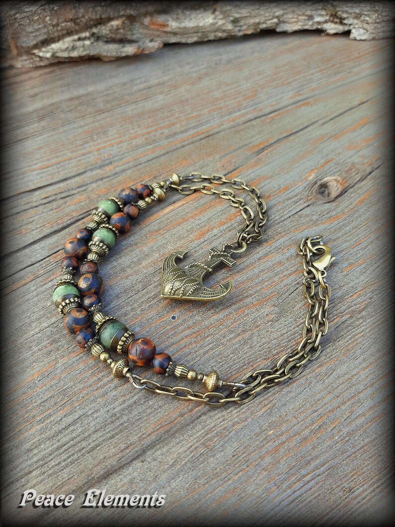 May include: A double-strand bracelet featuring bronze-toned metal chains and an anchor charm. The design incorporates round beads in mottled brown, blue, and green hues, accented with small, textured bronze-colored metal spacers throughout the piece.