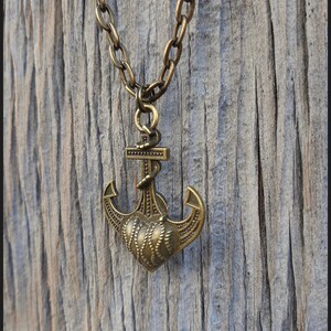May include: A bronze-toned metal necklace featuring a pendant shaped like a nautical anchor combined with a heart design. The pendant hangs from a matching thick oval-link chain. The text Peace Elements is visible in the bottom right corner.