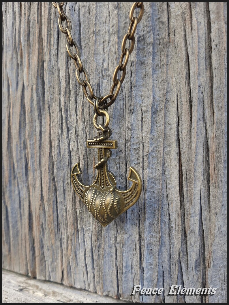 May include: A bronze-toned metal necklace featuring a pendant shaped like a nautical anchor combined with a heart design. The pendant hangs from a matching thick oval-link chain. The text Peace Elements is visible in the bottom right corner.