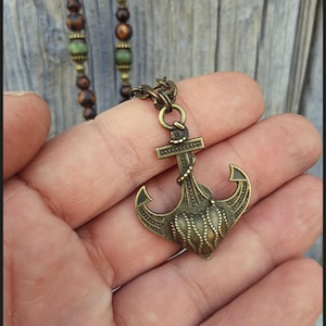 May include: A bronze-toned metal anchor pendant featuring a textured rope design on the fluke, suspended from a chain. The necklace includes a strand of brown and green beads. The text Peace Elements is visible in the bottom left corner.